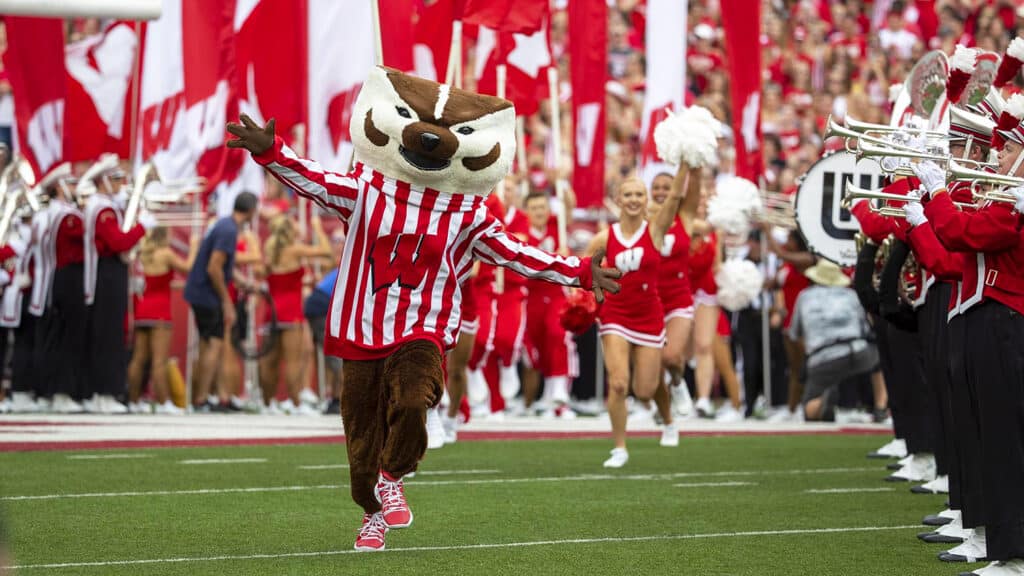 Bucky Badger Mascot running onto the football field with the band and cheerleaders.