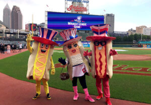 The Cleveland Guardians Racing Mascot posing for a photo on the Guardian's Field. On the left is the Ketchup Hotdog, middle is the Onion Hotdog, and on the right is the Mustard Hotdog.