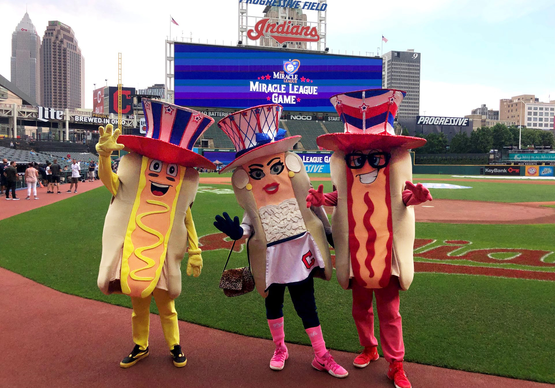 The Cleveland Guardians Racing Mascot posing for a photo on the Guardian's Field. On the left is the Ketchup Hotdog, middle is the Onion Hotdog, and on the right is the Mustard Hotdog.